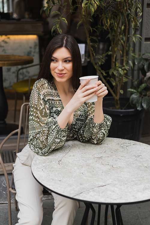 young woman with long brunette hair sitting on chair near round bistro table and holding paper cup with drink while looking away and smiling near blurred plants on terrace of cafe in Istanbul,stock image
