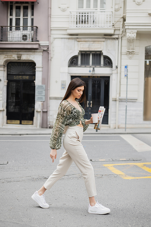 pretty young woman with long brunette hair and makeup holding paper cup with coffee and newspaper while walking in trendy outfit with beige pants and blouse on urban street near building in Istanbul,stock image