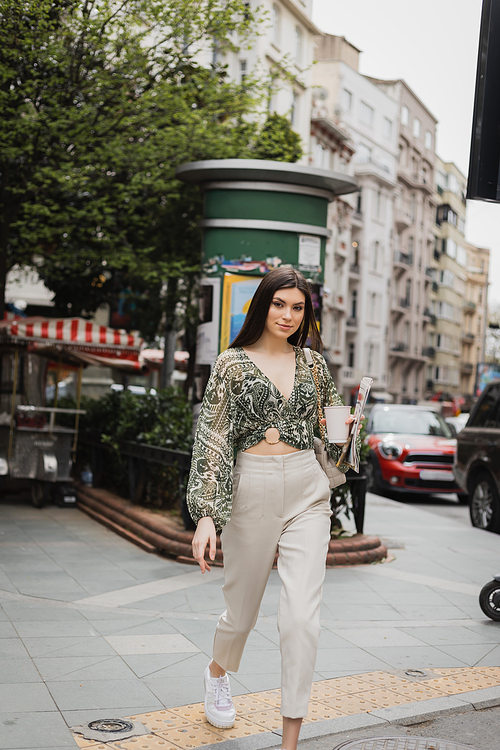 pretty woman with long hair and makeup holding paper cup with coffee and newspaper while walking in trendy outfit with handbag on chain strap on street near blurred building and cars in Istanbul,stock image