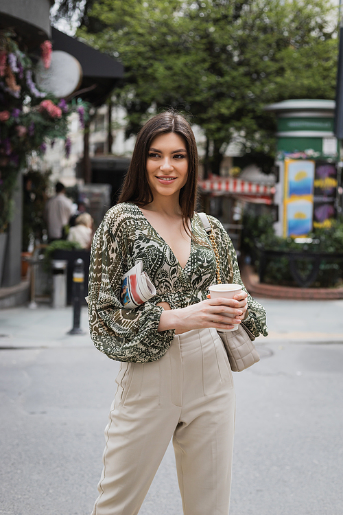 beautiful woman with long hair holding morning coffee in paper cup and newspaper while standing in trendy outfit with handbag and smiling on urban street near blurred flower shop in Istanbul,stock image