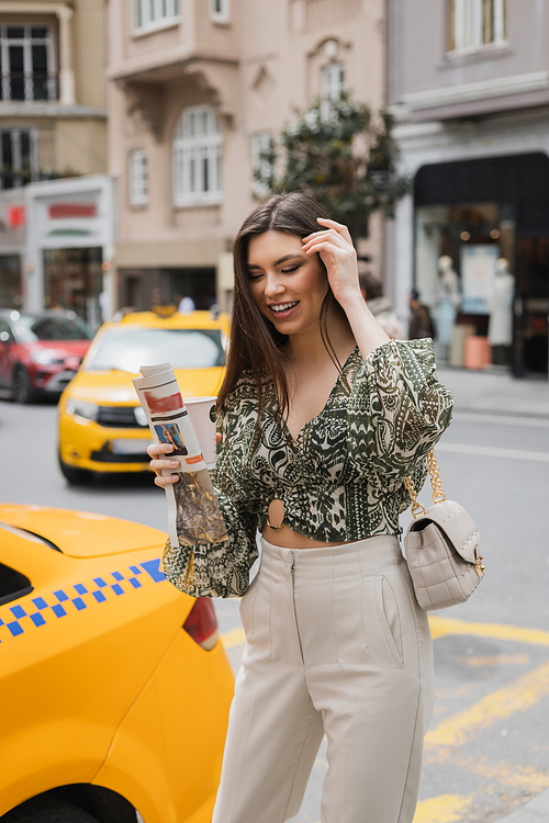 happy woman adjusting her long hair and holding paper cup with coffee and newspaper while standing in trendy outfit with handbag on chain strap near yellow taxi on blurred urban street in Istanbul,stock image