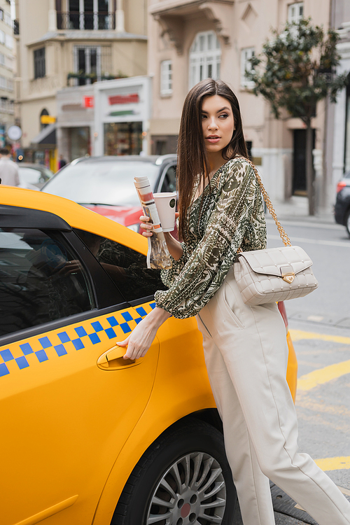 chic woman with long hair holding paper cup with coffee and newspaper while standing in trendy outfit with handbag on chain strap and opening door of yellow taxi on blurred urban street in Istanbul,stock image