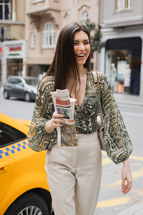 positive woman with long hair holding paper cup with coffee and newspaper while walking in trendy outfit with handbag on chain strap near yellow taxi on blurred urban street in Istanbul,stock image
