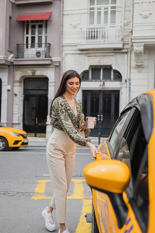 smiling young woman with long hair holding coffee in paper cup while standing in trendy outfit with handbag on chain strap and opening door of yellow cab on blurred urban street in Istanbul,stock image