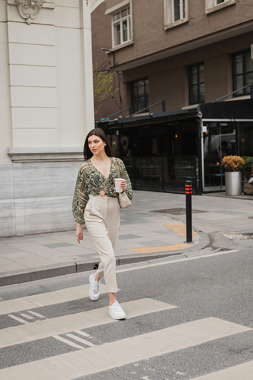 stylish woman with long hair in trendy outfit with beige pants, cropped blouse and handbag with chain strap walking on crosswalk with coffee on urban street with blurred building in Istanbul,stock image