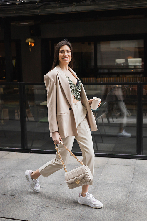 smiling woman with long hair walking in blazer with beige pants, cropped blouse and handbag on chain strap and holding paper cup with coffee near outdoor cafe on urban street in Istanbul,stock image