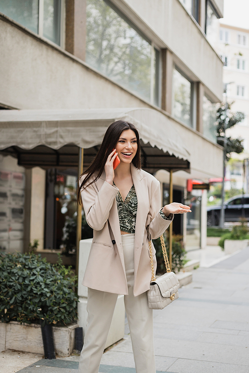 happy young woman with brunette long hair and makeup smiling while talking on smartphone and standing in trendy outfit with handbag on chain strap near blurred fancy restaurant in Istanbul,stock image
