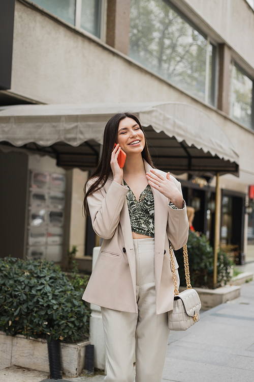 smiling young woman with brunette long hair and makeup smiling while talking on smartphone and standing in trendy outfit with handbag on chain strap near blurred restaurant in Istanbul,stock image