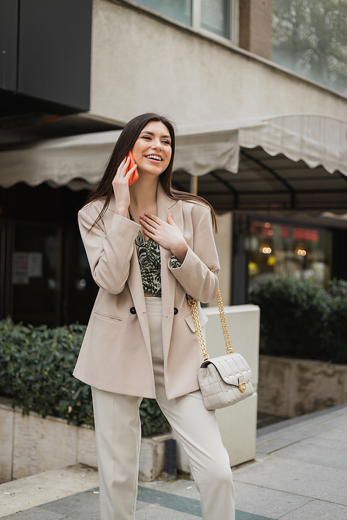 beautiful woman with brunette long hair and makeup smiling while talking on smartphone and standing in trendy outfit with handbag on chain strap near blurred fancy restaurant in Istanbul,stock image