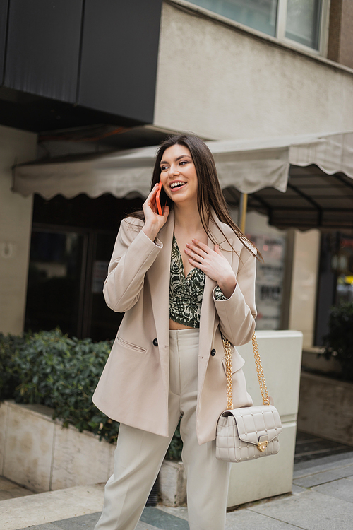 positive young woman with brunette long hair and makeup smiling while talking on smartphone and standing in trendy outfit with handbag on chain strap near blurred fancy restaurant in Istanbul,stock image