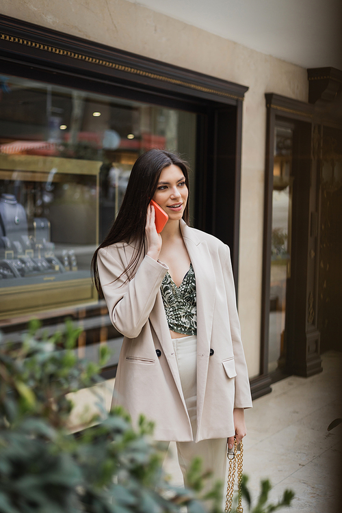 stylish young woman with brunette long hair and makeup talking on smartphone and standing in trendy outfit with handbag on chain strap near blurred jewelry store and plant in Istanbul,stock image