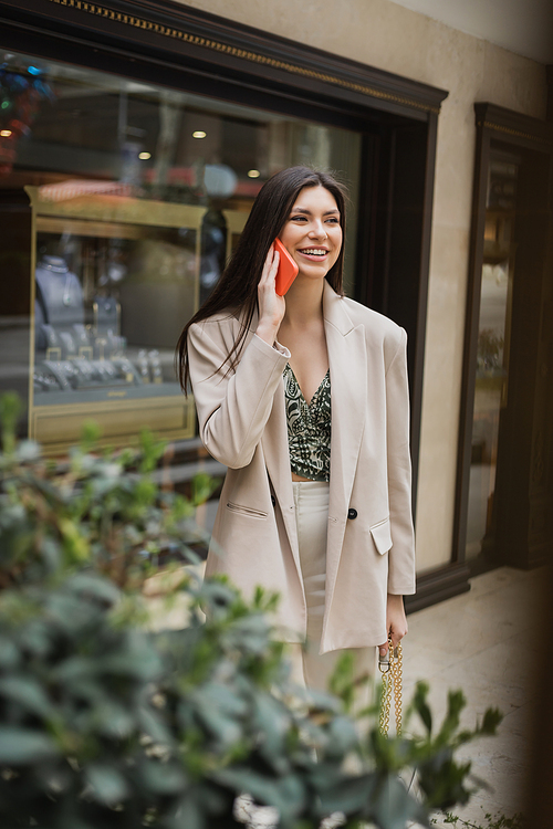 smiling young woman with brunette long hair and makeup talking on smartphone and standing in trendy outfit with handbag on chain strap near blurred jewelry store and plant in Istanbul,stock image