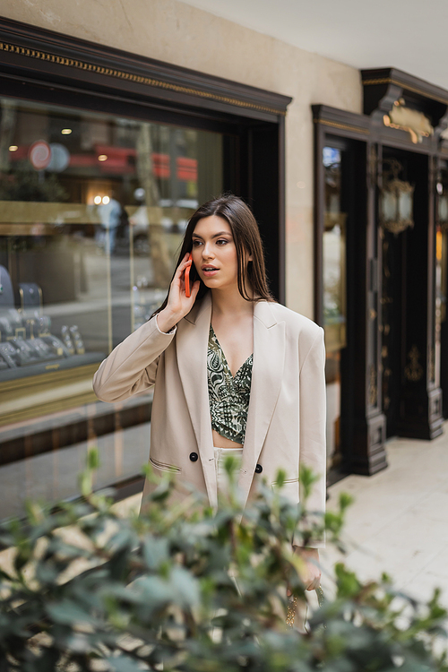 surprised young woman with brunette long hair and makeup talking on smartphone and standing in trendy outfit with handbag on chain strap near blurred jewelry store and plant in Istanbul,stock image