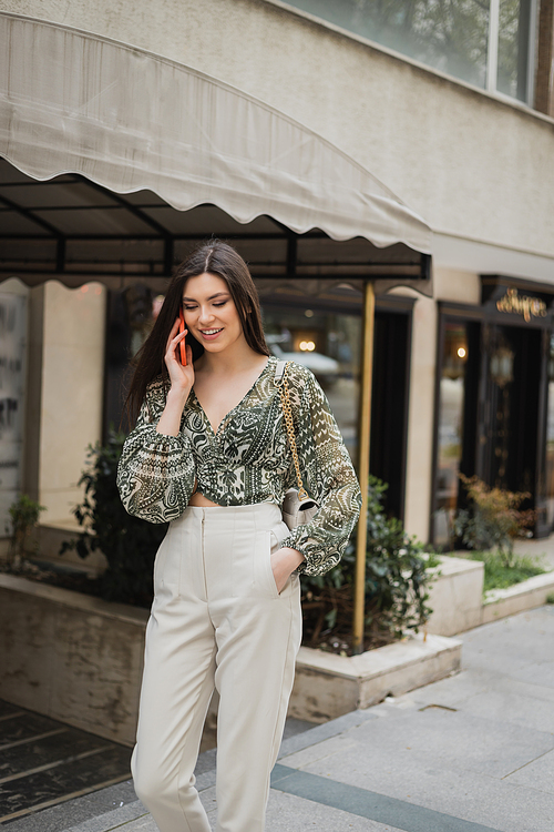 stylish young woman with brunette long hair and makeup smiling while talking on smartphone and standing in trendy outfit with handbag on chain strap near blurred fancy restaurant in Istanbul,stock image