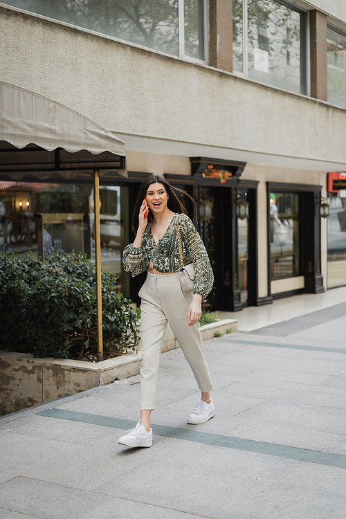 amazed young woman with brunette long hair and makeup smiling while talking on smartphone and walking in trendy outfit with handbag on chain strap near blurred fancy restaurant in Istanbul,stock image