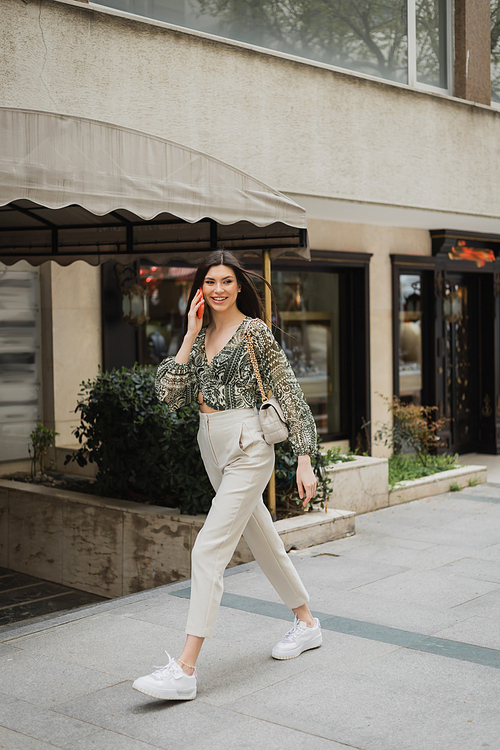 cheerful young woman with brunette long hair and makeup smiling while talking on smartphone and walking in trendy outfit with handbag on chain strap near blurred fancy restaurant in Istanbul,stock image