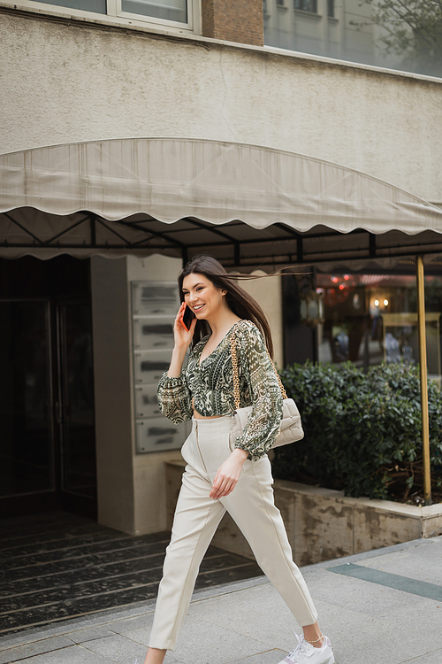 cheerful young woman with long hair and makeup smiling while talking on smartphone and walking in trendy outfit with handbag on chain strap near blurred fancy restaurant in Istanbul,stock image