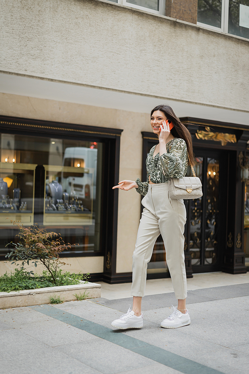 cheerful young woman with long hair and makeup smiling while talking on smartphone and walking in trendy outfit with handbag on chain strap near blurred jewelry store in Istanbul,stock image