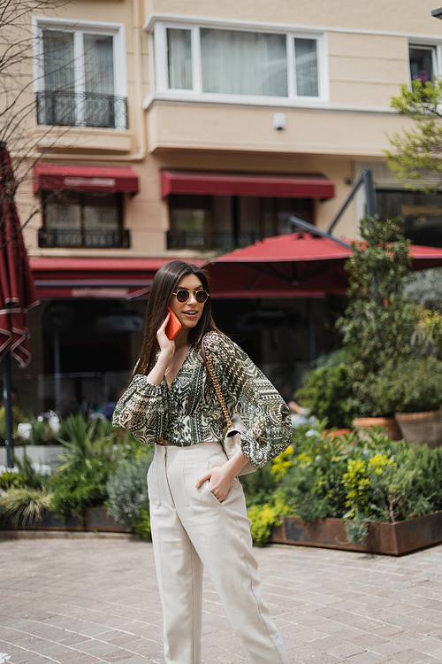 young woman with long hair and sunglasses smiling while talking on smartphone and standing with hand in pocket and handbag on chain strap near blurred building and plants in Istanbul,stock image