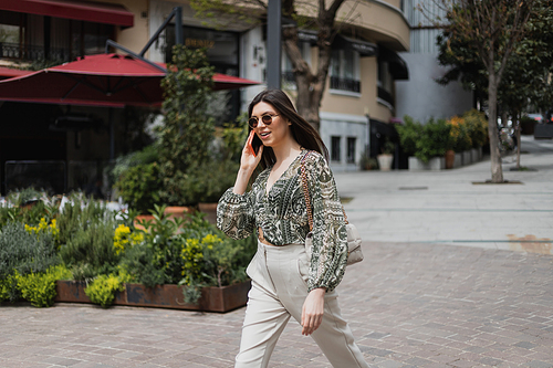 cheerful young woman with long hair and sunglasses smiling while talking on smartphone and walking in trendy outfit with handbag on chain strap near blurred building and plants on street in Istanbul,stock image