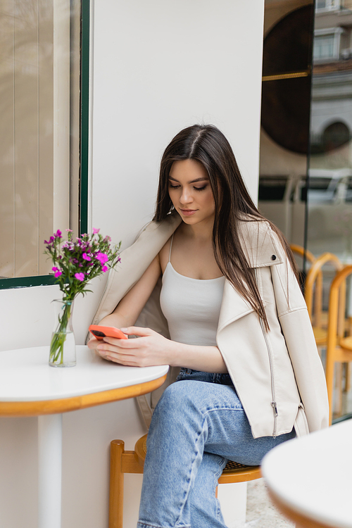 young woman with long hair sitting on chair near bistro table with flowers in vase and texting on smartphone while sitting in trendy clothes with leather jacket in cafe on terrace outdoors in Istanbul,stock image