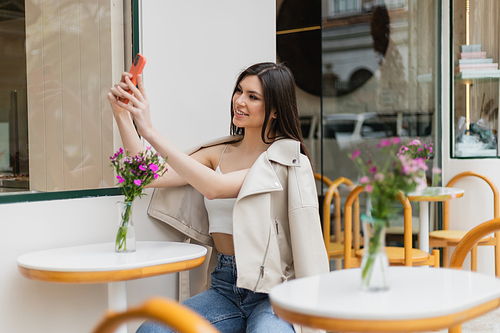 happy woman with long hair sitting on chair near bistro table with flowers in vase and taking selfie on smartphone while posing in trendy clothes in cafe on terrace outdoors in Istanbul,stock image