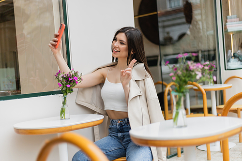 happy woman with long hair sitting on chair near bistro table with flowers in vase and taking selfie, shoving v sign, posing in trendy clothes in cafe on terrace outdoors in Istanbul,stock image
