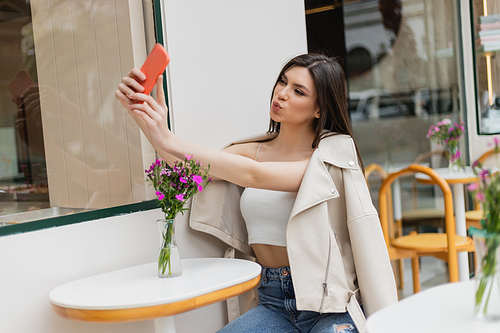 woman with long hair sitting on chair near bistro table with flowers in vase and pouting lips, taking selfie on smartphone while posing in trendy clothes in cafe on terrace outdoors in Istanbul,stock image