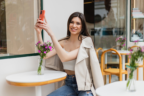 smiling woman with long hair sitting on chair near bistro table with flowers in vase and taking selfie on smartphone while posing in trendy clothes in cafe on terrace outdoors in Istanbul,stock image