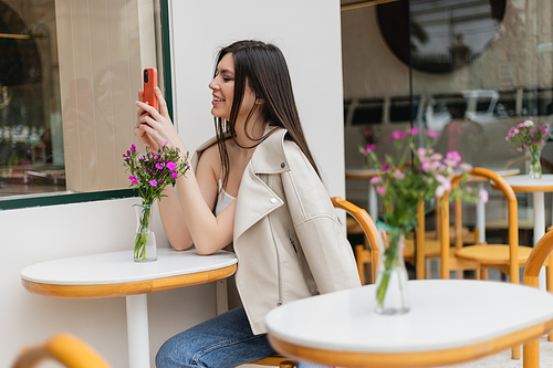 happy woman with long hair sitting on chair near bistro table with flowers in vase and messaging on smartphone while sitting in trendy clothes in cafe on terrace outdoors in Istanbul,stock image