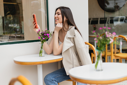 happy woman with long hair sitting on chair near bistro table with flowers in vase and texting on smartphone while sitting in trendy clothes with beige blazer in cafe on terrace outdoors in Istanbul,stock image