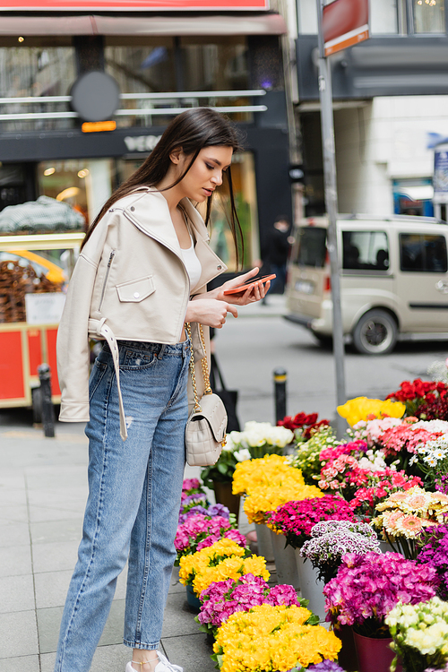 brunette woman with long hair standing in beige leather jacket, denim jeans and handbag with chain strap holding smartphone while looking at flowers near blurred cars on street in Istanbul, vendor,stock image