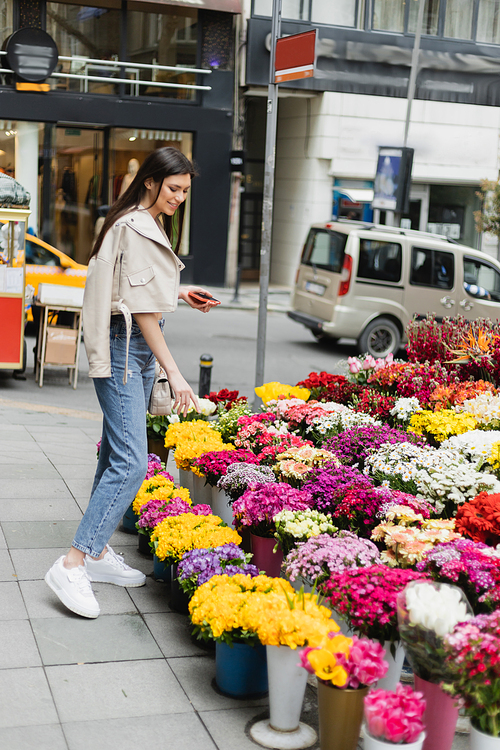 happy woman with long hair standing in beige leather jacket, denim jeans and handbag with chain strap, holding smartphone near bouquet of flowers next to blurred cars on street in Istanbul, vendor,stock image