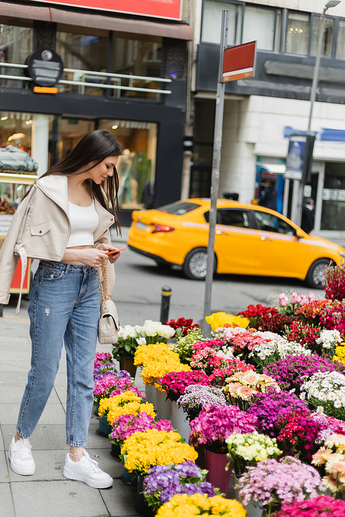 brunette woman with long hair standing in beige leather jacket, denim jeans and handbag with chain strap holding smartphone near flowers next to blurred street in Istanbul, vendor,stock image