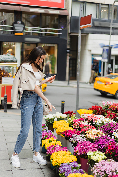 brunette woman with long hair standing in beige leather jacket and denim jeans while holding smartphone and pointing at bouquets of flowers next to blurred car on street in Istanbul, vendor,stock image
