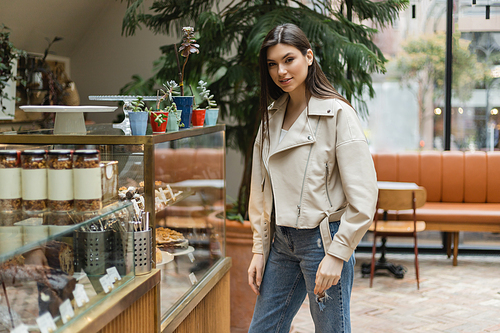 brunette young woman with long hair in beige leather jacket and denim jeans looking at camera while standing near cake display with pastry and jars of jam in modern bakery shop in Istanbul,stock image