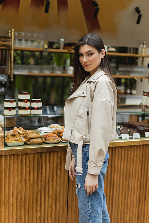 brunette young woman with long hair in beige leather jacket and denim jeans looking at camera while standing near cake display with tasty pastry and jars of jam in modern bakery shop in Istanbul,stock image