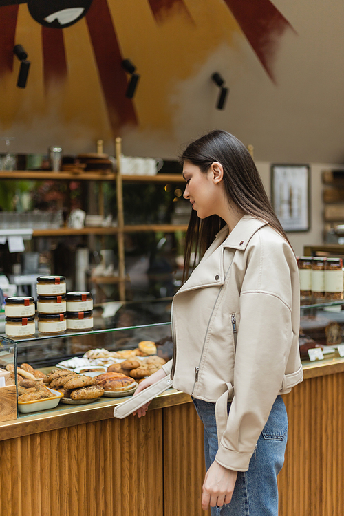 young woman with brunette long hair in beige leather jacket and denim jeans standing near cake display with delicious pastry and jars of jam in modern bakery shop in Istanbul,stock image