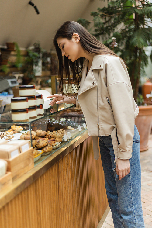 brunette young woman with long hair in beige leather jacket and denim jeans standing near cake display and choosing pastry near jars of jam in modern bakery shop in Istanbul,stock image
