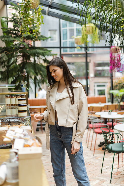 brunette young woman with long hair in beige leather jacket and denim jeans standing near cake display with pastry and jars of jam in modern bakery shop in Istanbul,stock image