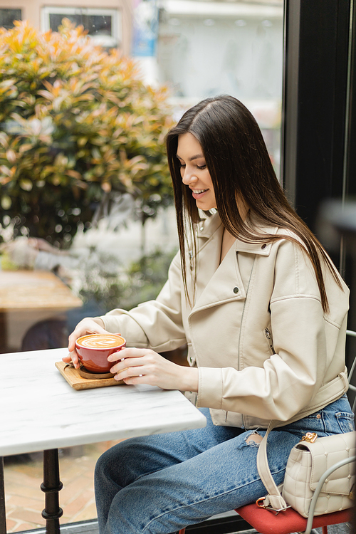 happy brunette woman in leather jacket sitting on chair next to window and bistro table while holding cup of cappuccino with coffee art inside of modern cafe in Istanbul,stock image