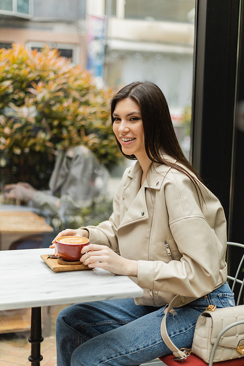 happy brunette woman in leather jacket sitting on chair next to window and bistro table while holding cup of delicious cappuccino with coffee art inside of modern cafe in Istanbul,stock image