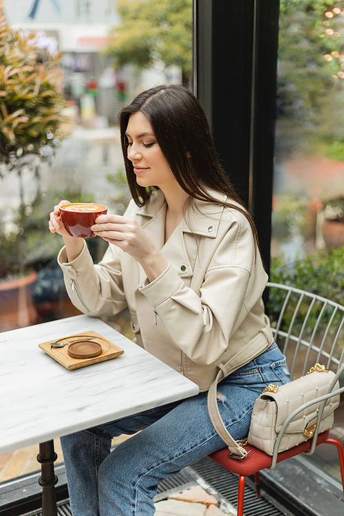 happy brunette woman in leather jacket sitting on chair next to window and bistro table while holding cup of latte with foam and coffee art inside of modern cafe in Istanbul,stock image