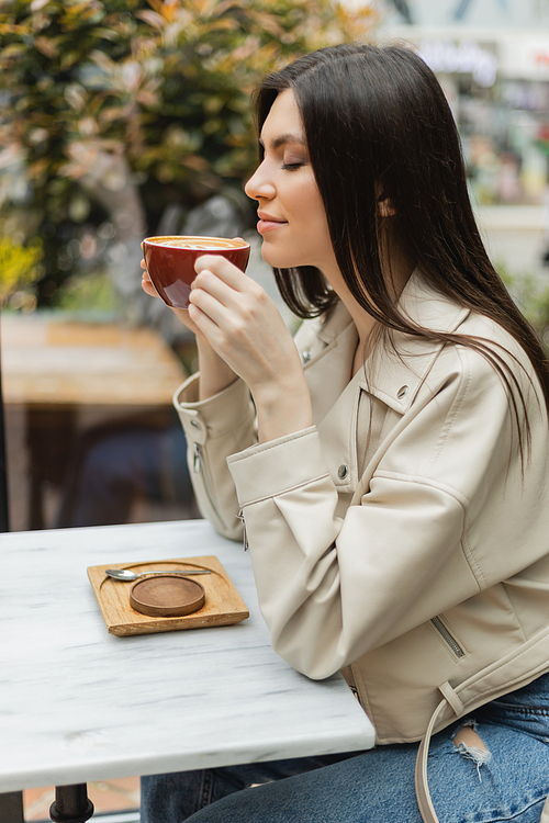 happy brunette woman with closed eyes in leather jacket sitting on chair next to window and bistro table while holding cup of cappuccino inside of modern cafe in Istanbul,stock image