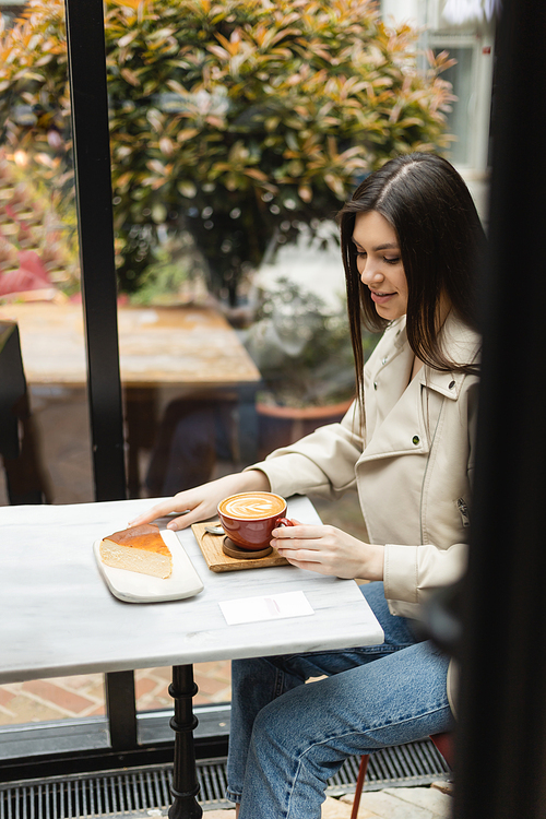 happy woman with long hair sitting in leather jacket next to window and bistro table while holding cup of cappuccino with coffee art and looking at cheesecake inside of modern cafe in Istanbul,stock image