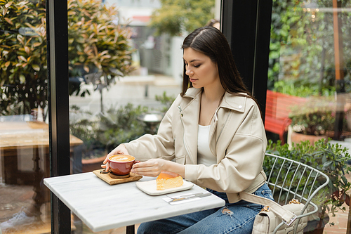 brunette woman with long hair sitting in leather jacket next to window and bistro table while holding cup of cappuccino with coffee art near cheesecake inside of modern cafe in Istanbul,stock image