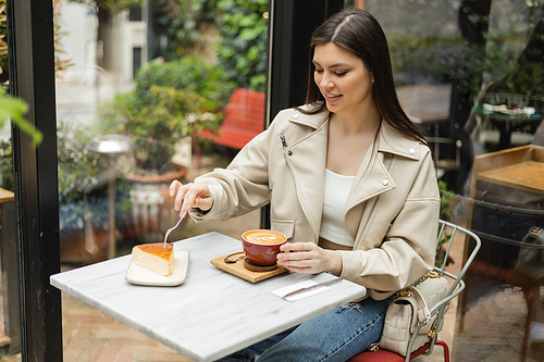 happy woman with long hair sitting in leather jacket next to window and bistro table while holding cup of cappuccino and fork above cheesecake inside of modern cafe in Istanbul,stock image