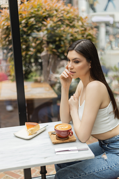 young woman with long hair sitting next to window and bistro table with cup of cappuccino and tasty cheesecake on plate while looking away inside of modern cafe in Istanbul,stock image