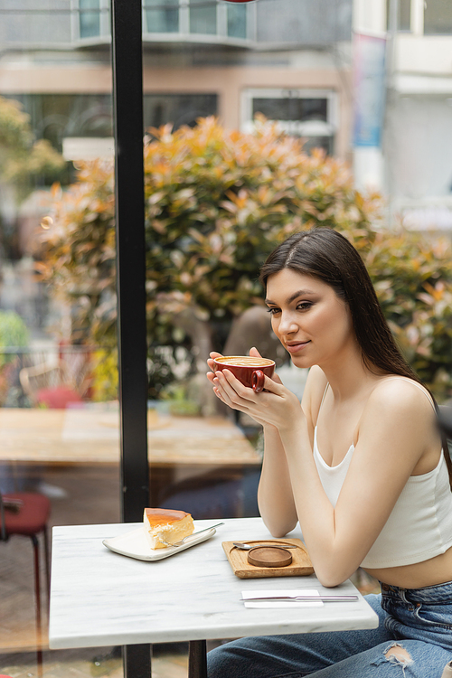 happy woman with long hair sitting next to window and holding cup of cappuccino with coffee art near cheesecake on plate of bistro table inside of modern cafe in Istanbul,stock image