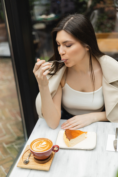 young woman with long hair eating cheesecake next to cup of cappuccino on bistro table while sitting in leather jacket next to window inside of modern cafe in Istanbul,stock image